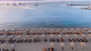 Une plage bordée de rangées de chaises longues et de parasols, une jetée avec des bateaux, et une mer calme.