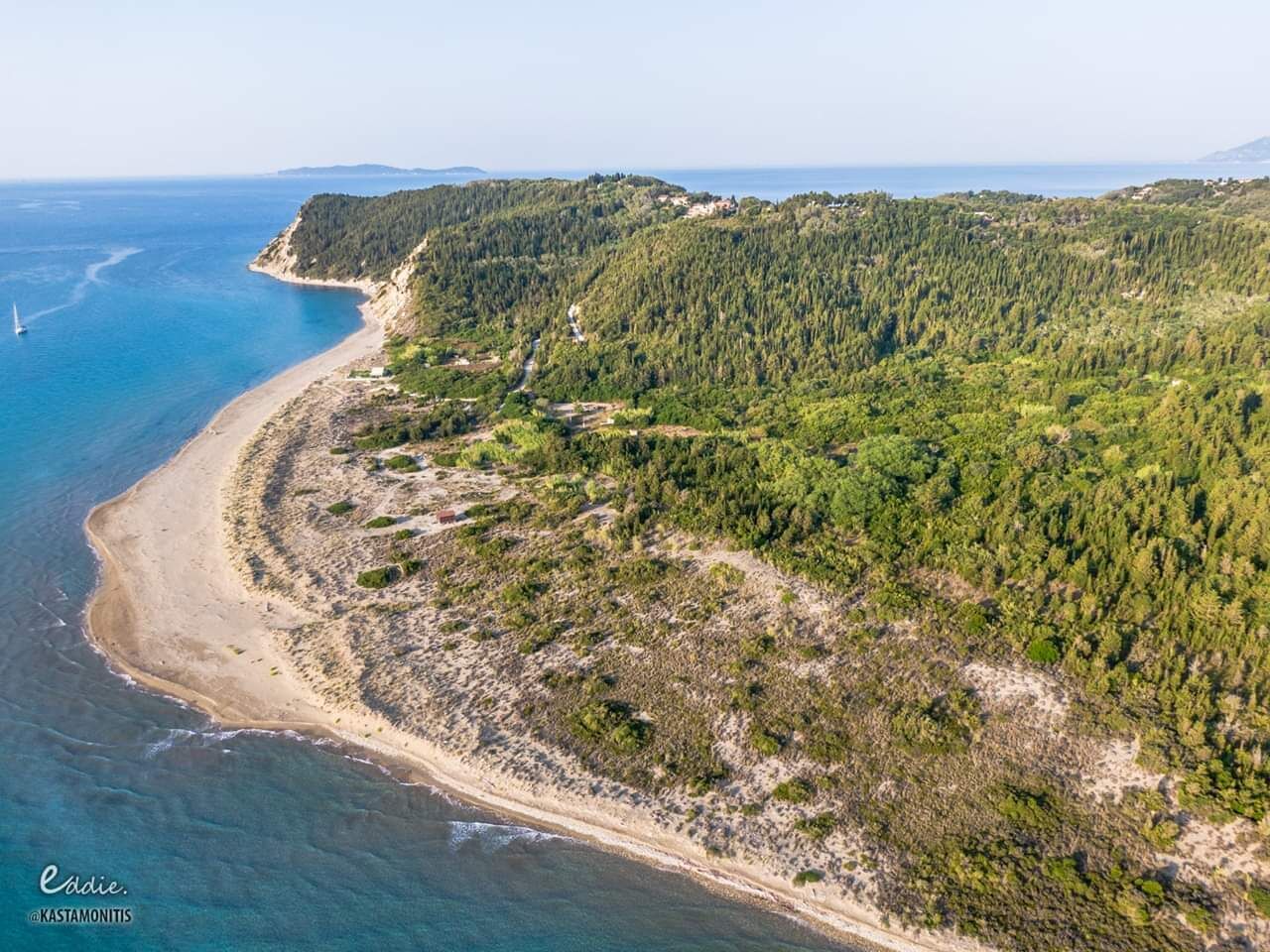 An aerial view of a coastal area with a sandy beach, dense forested hills, and a clear blue sea.
