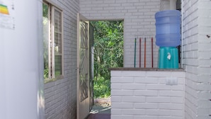 Un couloir étroit aux murs en briques blanches, un tabouret en bois, un distributeur d’eau bleu et une fenêtre avec des stores.
