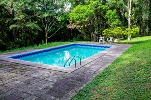 A rectangular swimming pool with a blue border, surrounded by a paved deck and lush greenery. - Chalet em Viamão (Viamao)