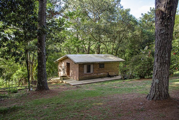 A small, rustic cabin with a brick exterior and a sloped roof, surrounded by dense trees and a grassy area. - Chalet em Viamão (Viamao)