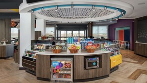 A modern food court with a central island, refrigerated display cases, and a seating area with tables and chairs. - Tru by Hilton Lubbock Southwest (Lubbock)