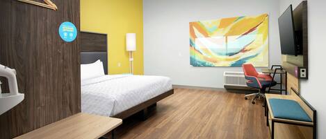 A hotel room with a bed, a desk, a chair, a TV, and a painting on the wall.