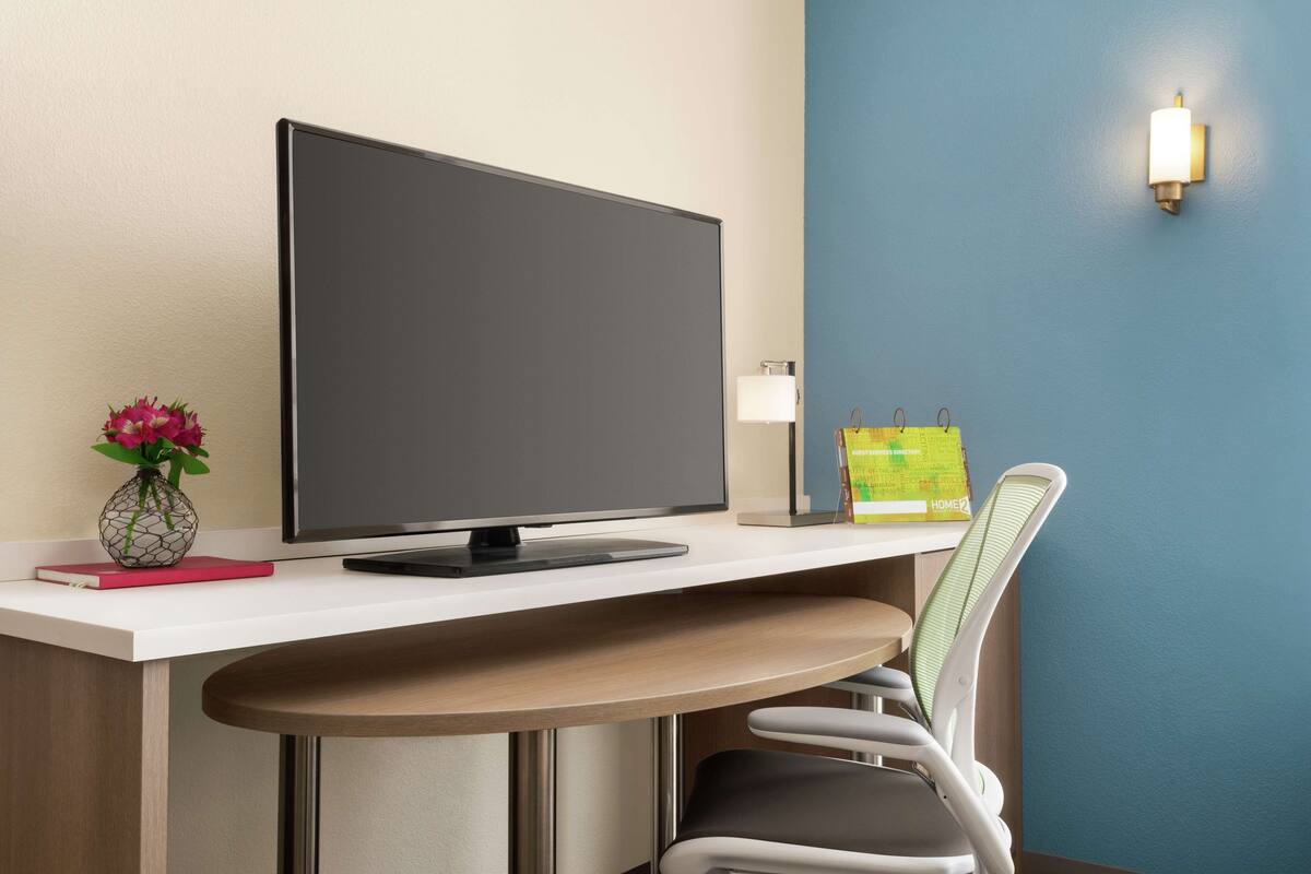 Een moderne hotelkamer met een flatscreen-tv, een bureau met een laptop, een vaas met bloemen, een stoel en een wandlamp.