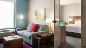A hotel room with a sofa, a small table, a lamp, a bed, and a painting on the wall. - Home2 Suites by Hilton Overland Park (Overland Park)