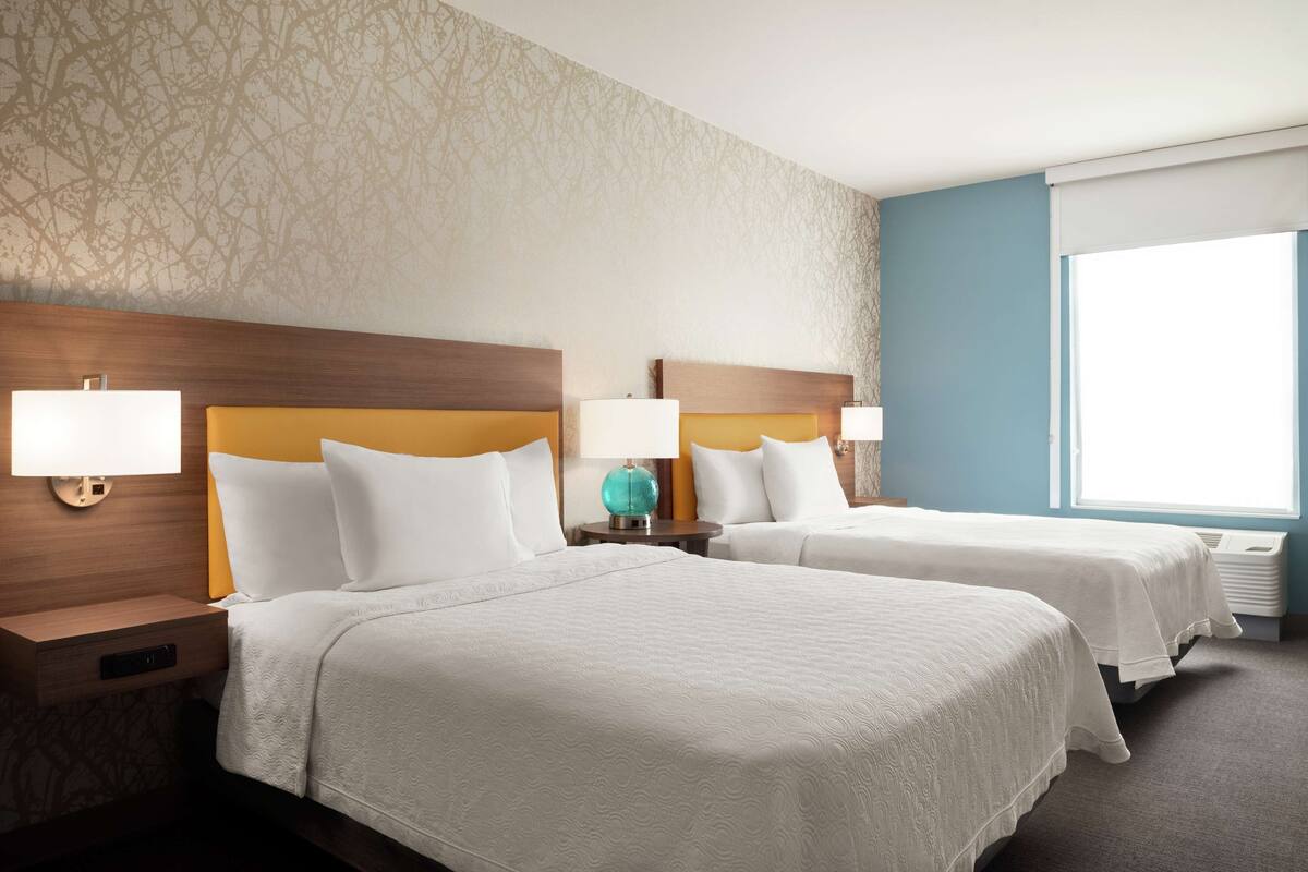 Hotelkamer met twee bedden, elk met een houten hoofdbord, wit beddengoed en bedlampjes. Er is een raam met jaloezieën en een kamer met een patroon.
