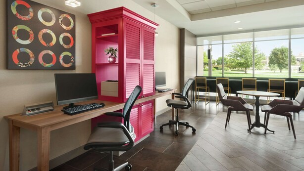 A modern office space with a wooden desk, a computer, and office chairs. There is a large window with a view of greenery outside. - Home2 Suites by Hilton Overland Park (Overland Park)
