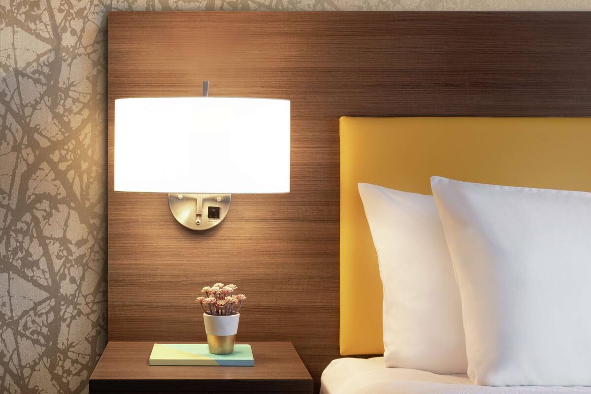 Een netjes opgemaakt bed met witte kussens, een nachtkastje met een lamp en een kleine vaas met bloemen.