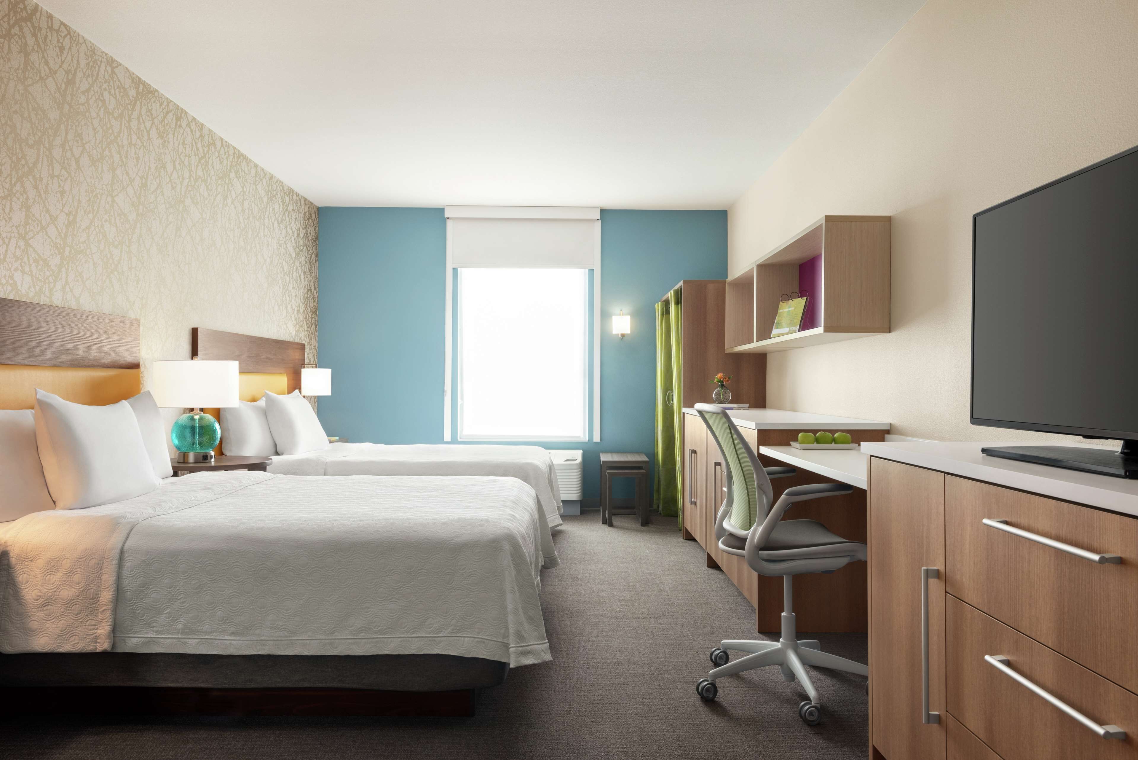 Une chambre d’hôtel avec deux lits, un bureau avec un ordinateur, une télévision et une fenêtre avec des stores.