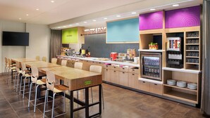 A modern kitchen area with a long wooden table, multiple chairs, a refrigerator, and a coffee machine. - Home2 Suites by Hilton Overland Park (Overland Park)