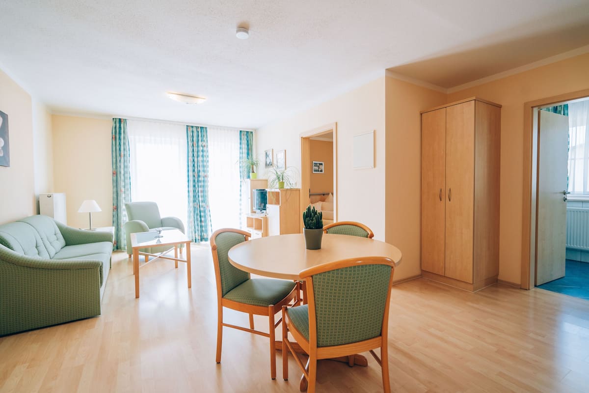 A modern living room with a round wooden table, green upholstered chairs, a sofa, and a large wardrobe.
