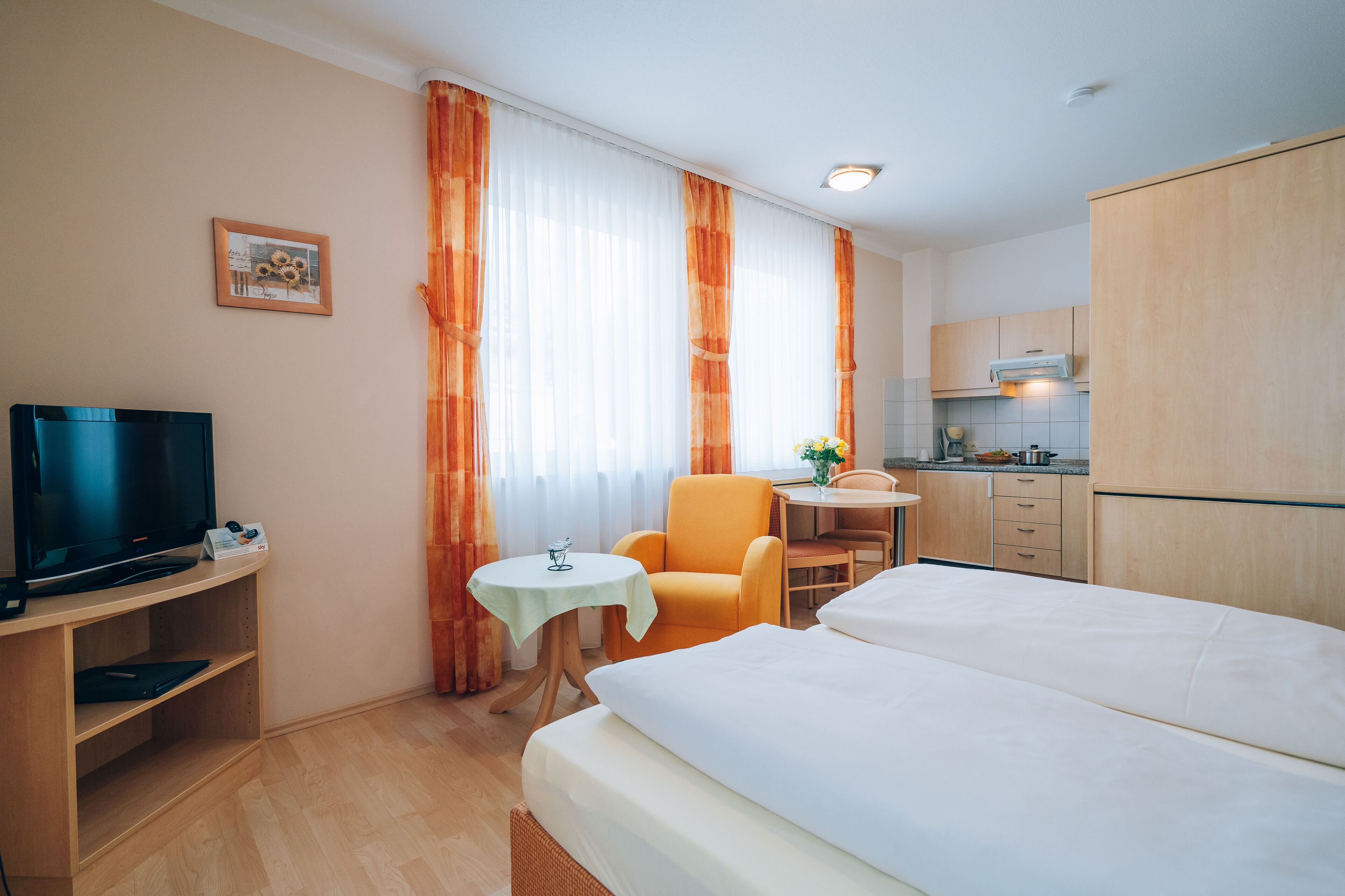 A hotel room with a bed, a desk with a TV, a small table, an orange chair, and a kitchenette.
