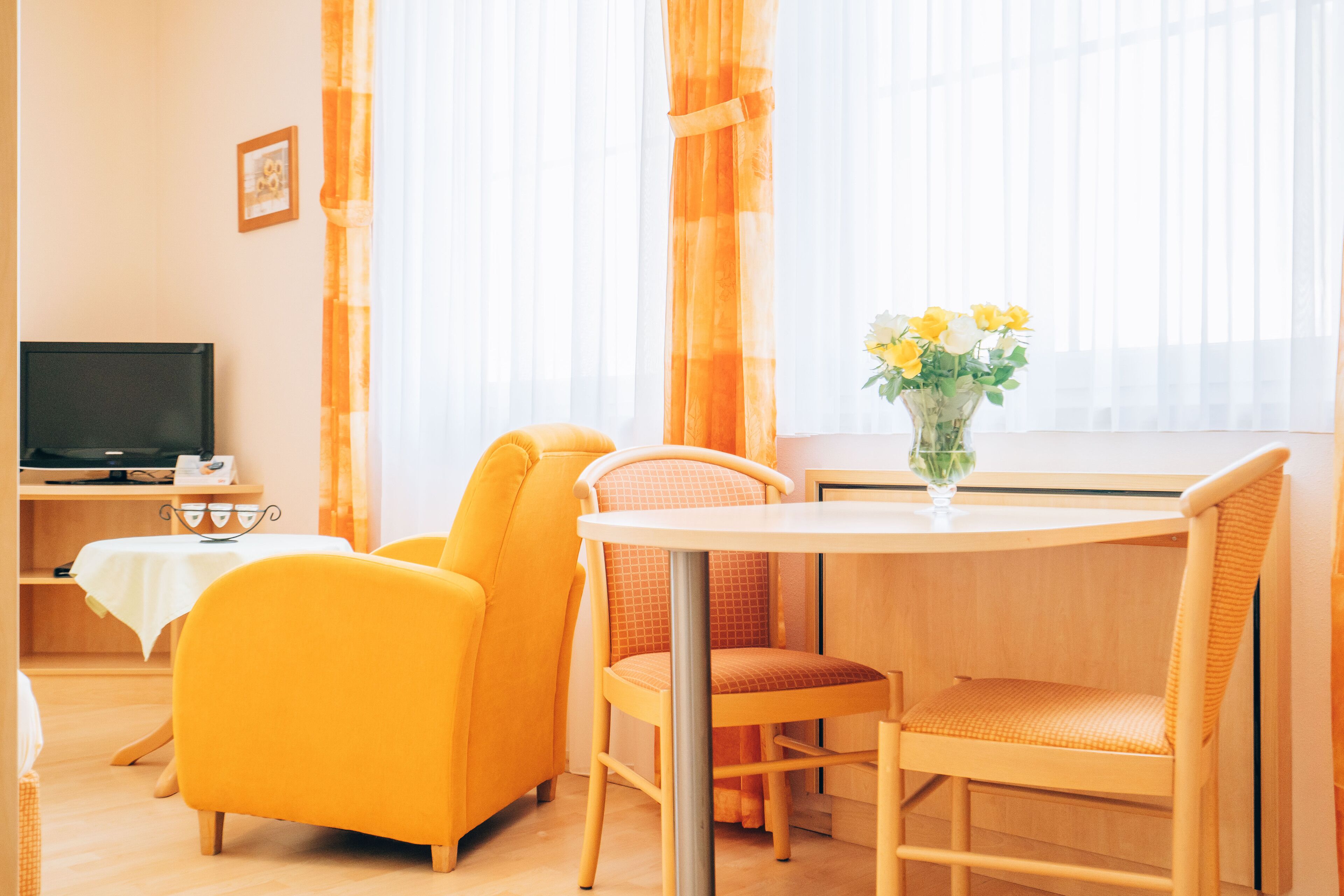 Una camera d'albergo con televisione, un tavolo da pranzo con sedie, un vaso con fiori e ampie finestre con tende.