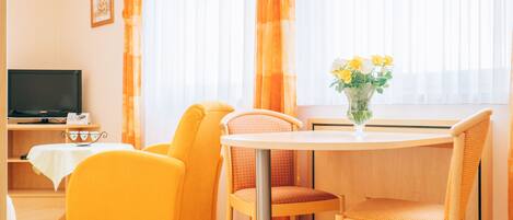 A hotel room with a television, a dining table with chairs, a vase with flowers, and large windows with curtains.