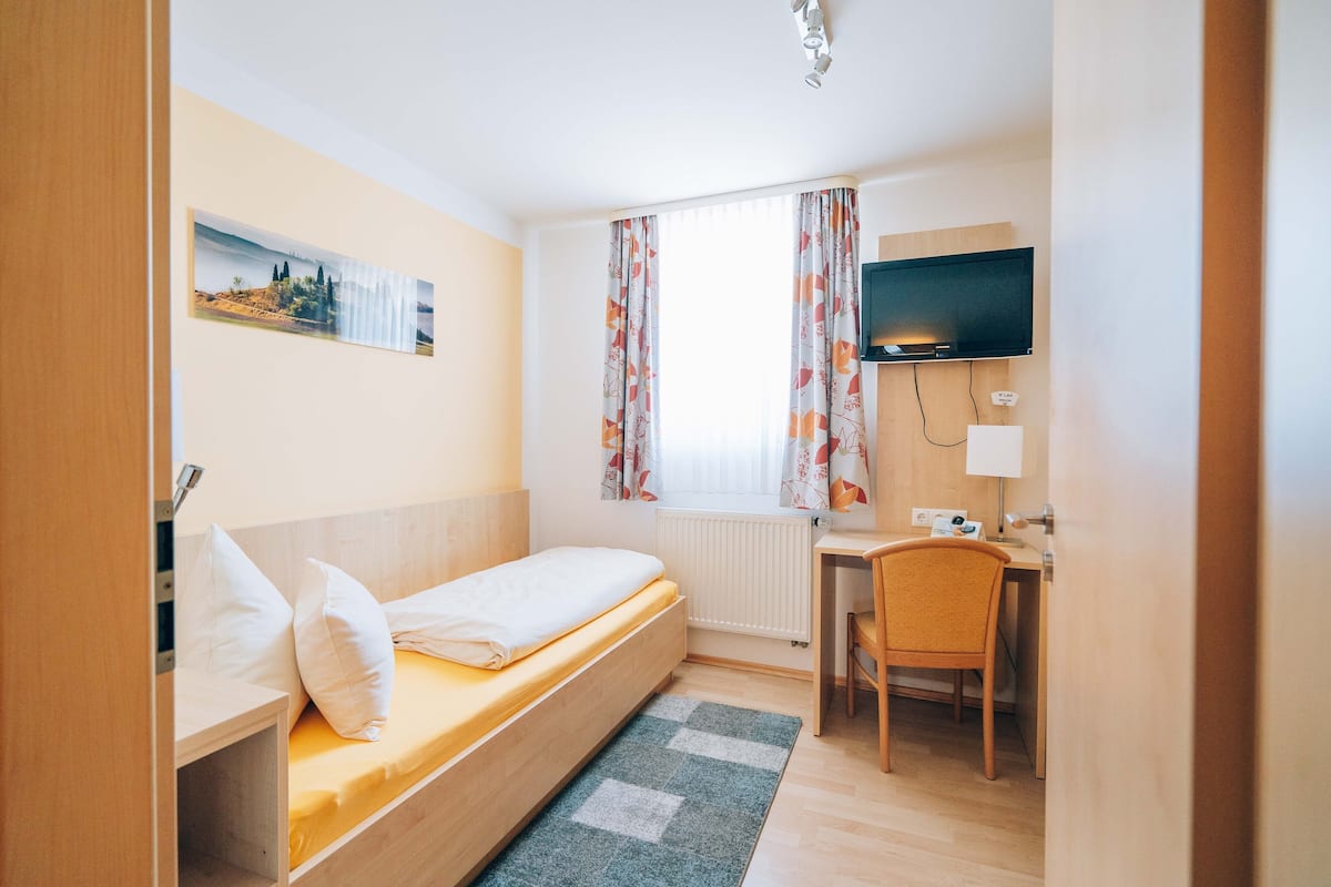 A compact hotel room with a single bed, a desk with a chair, a television, and a window with curtains.