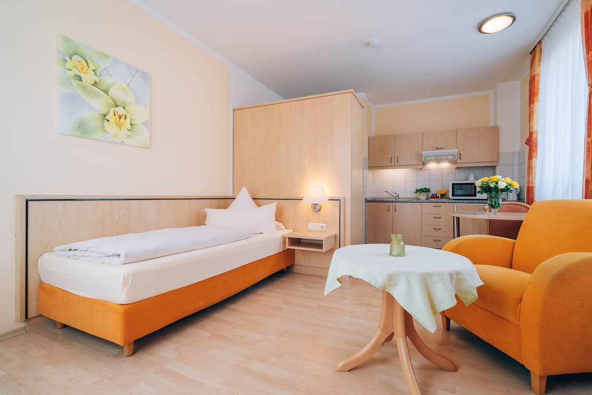 A hotel room with a bed, a small table, a sofa, a kitchen area, and a painting on the wall.