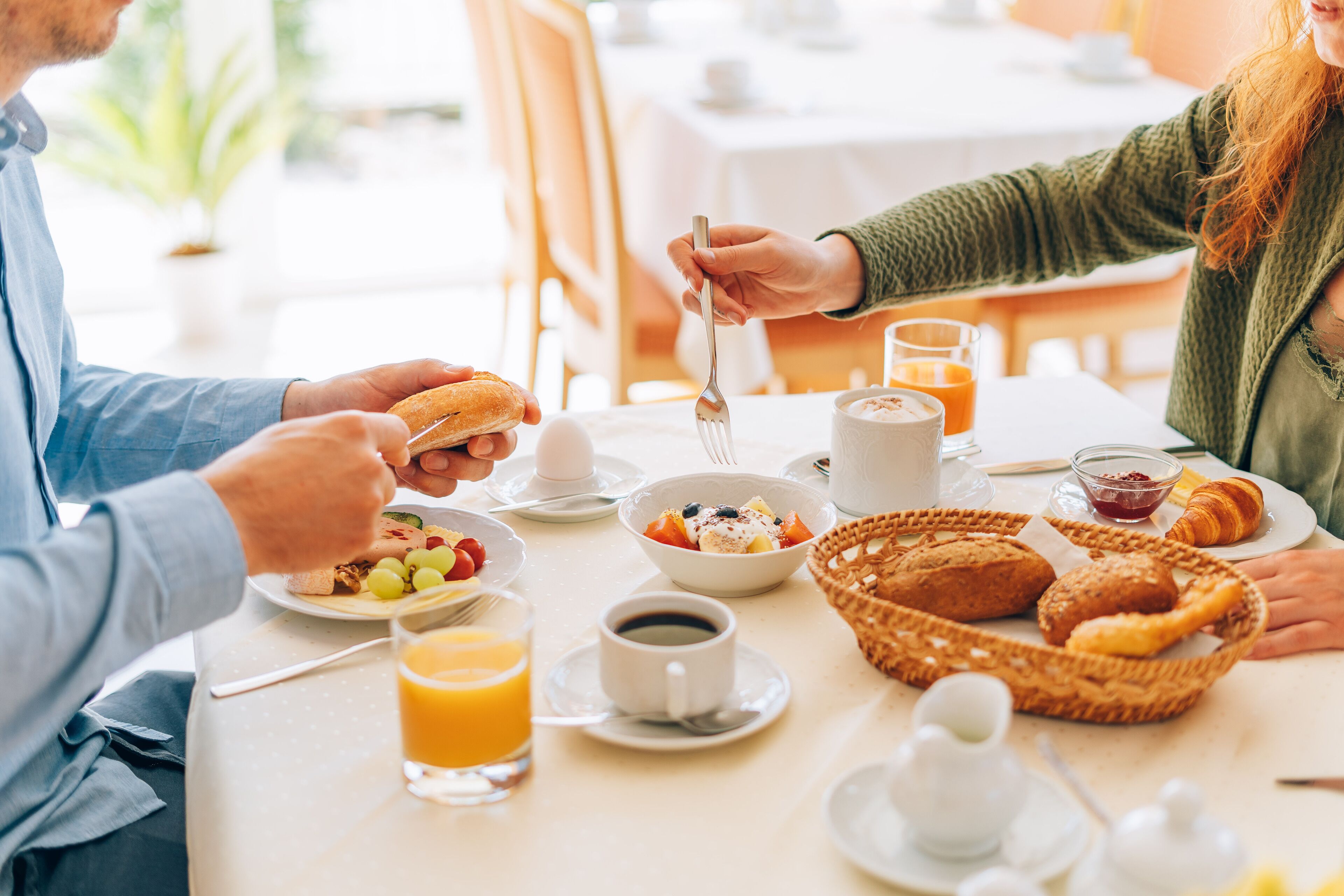 A breakfast setting with a table set for two, including plates with food, a basket of pastries, a glass of orange juice, and a cup of coffee.