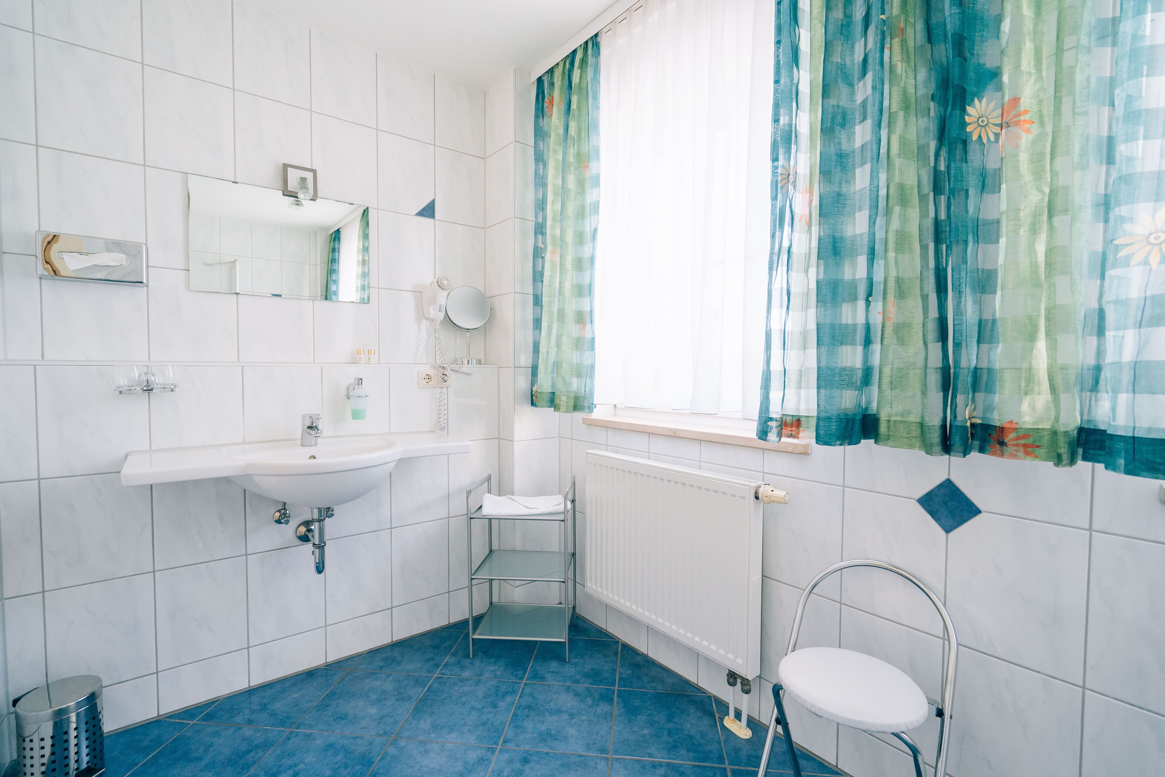 Un baño con lavamanos blanco, espejo y retrete, suelo de azulejos azules y una ventana con cortinas estampadas.