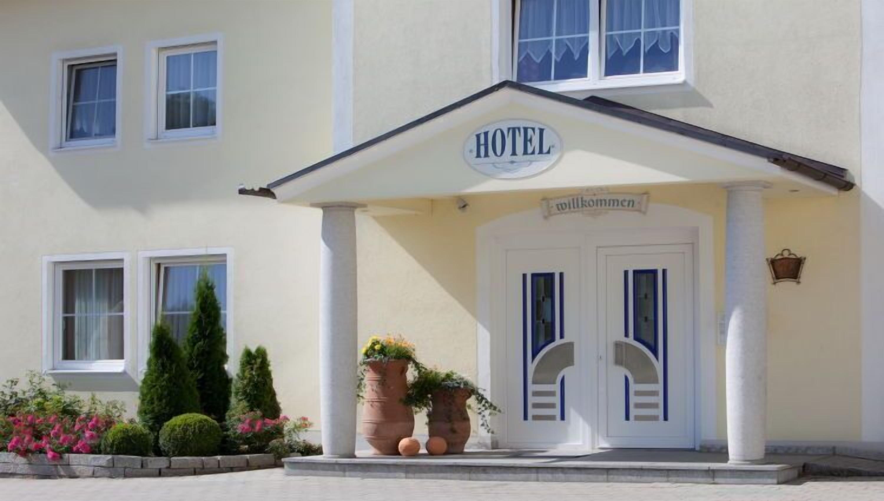 A hotel with a sign reading "HOTEL" above the entrance, a "Willkommen" sign, and a symmetrical facade with two doors and windows.