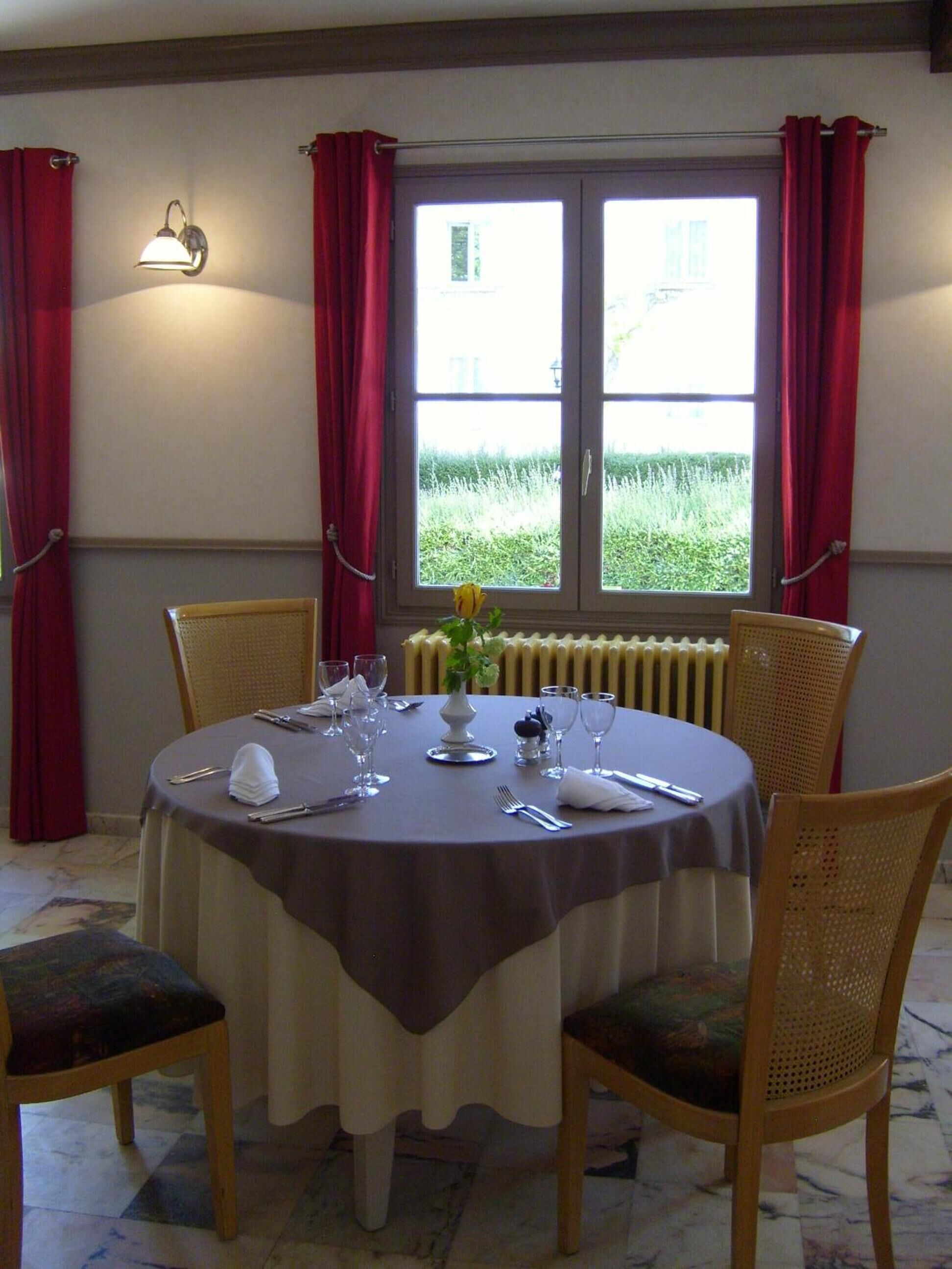 A dining table set for four with a vase of flowers, red curtains, and a window with a view of greenery.