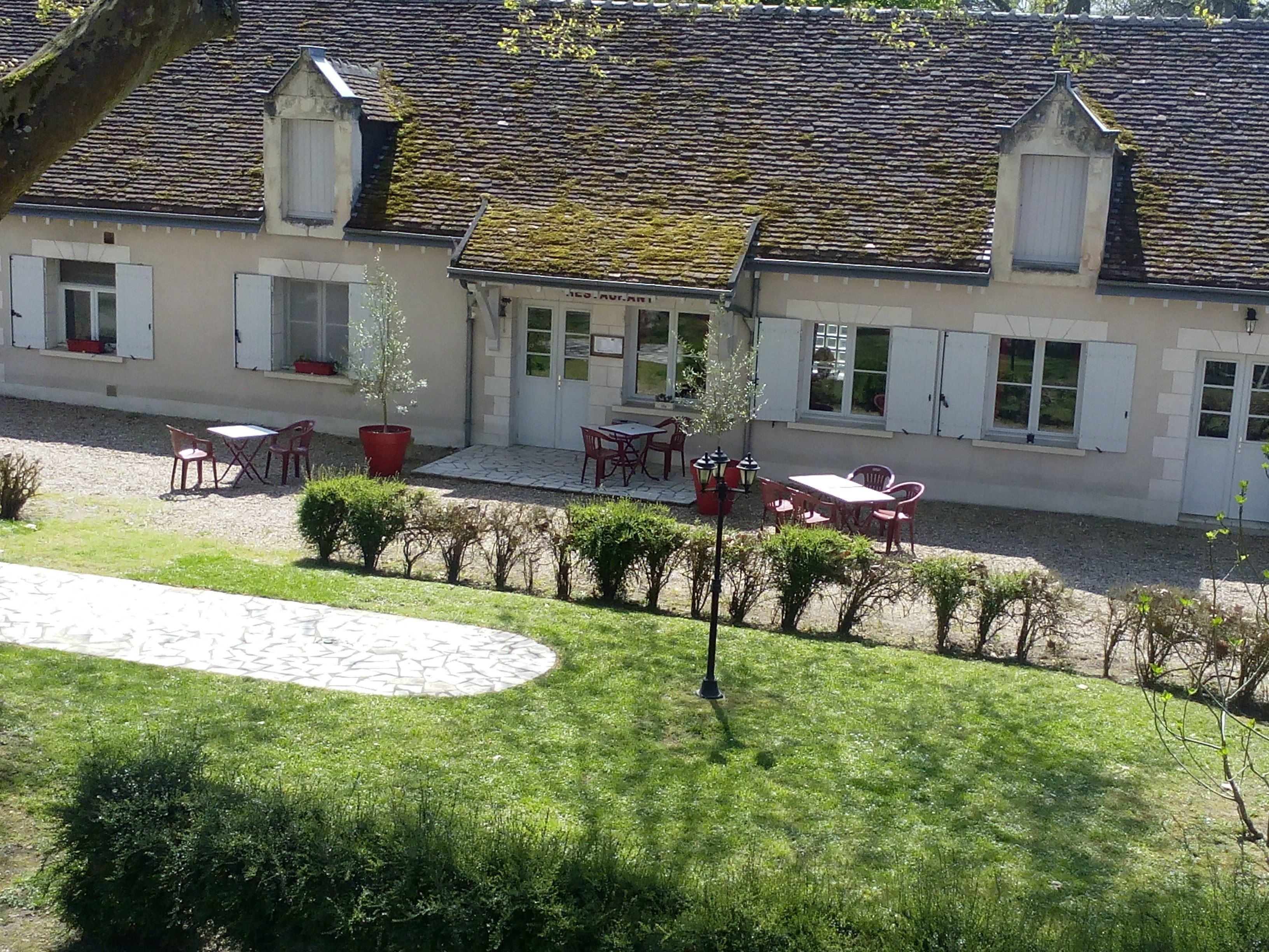 A building with a tiled roof, white exterior walls, and multiple windows. There is a paved area with outdoor furniture and a small garden.