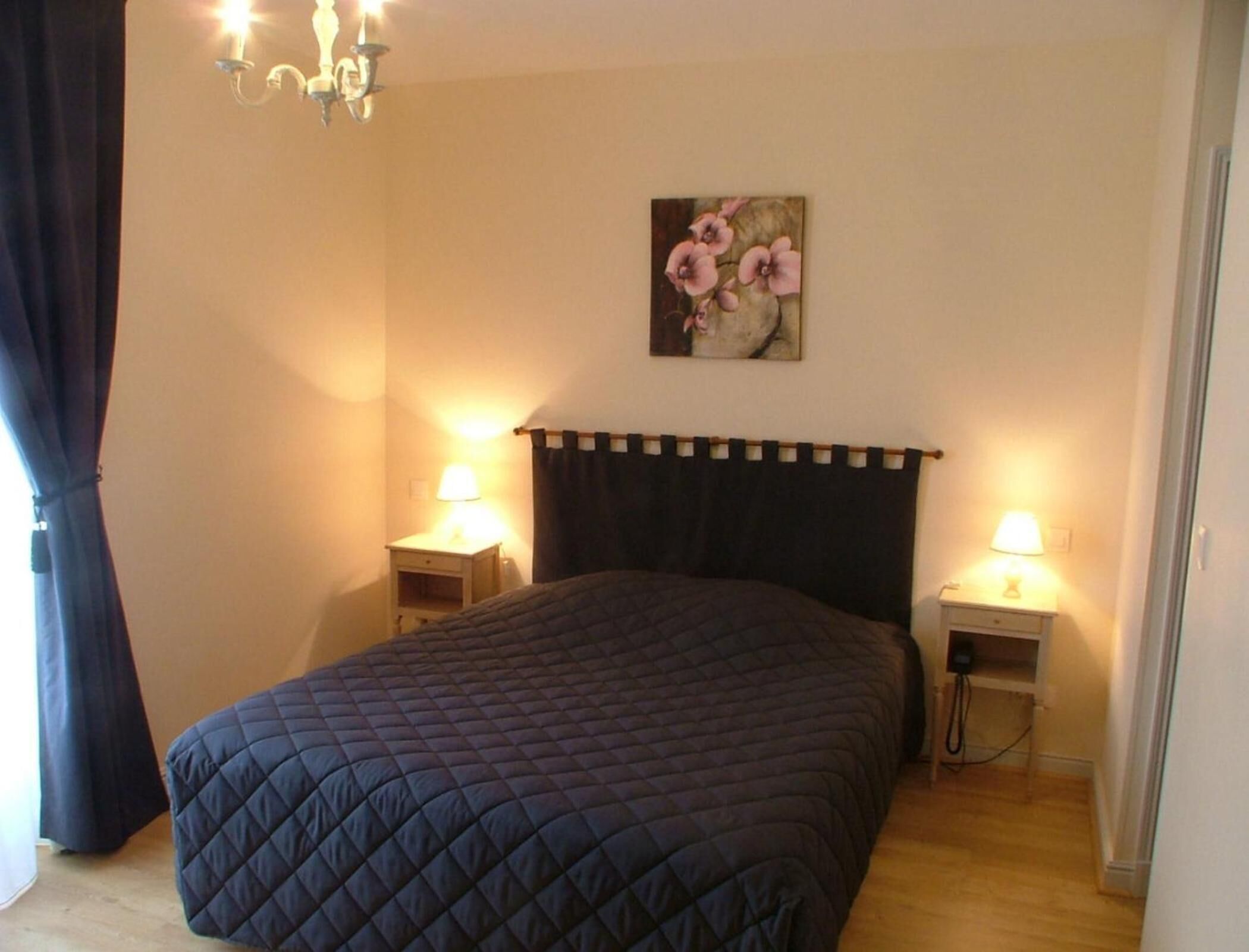 A bedroom with a dark quilted bedspread, a black headboard, two bedside lamps, a wall-mounted picture of flowers, and a chandelier.
