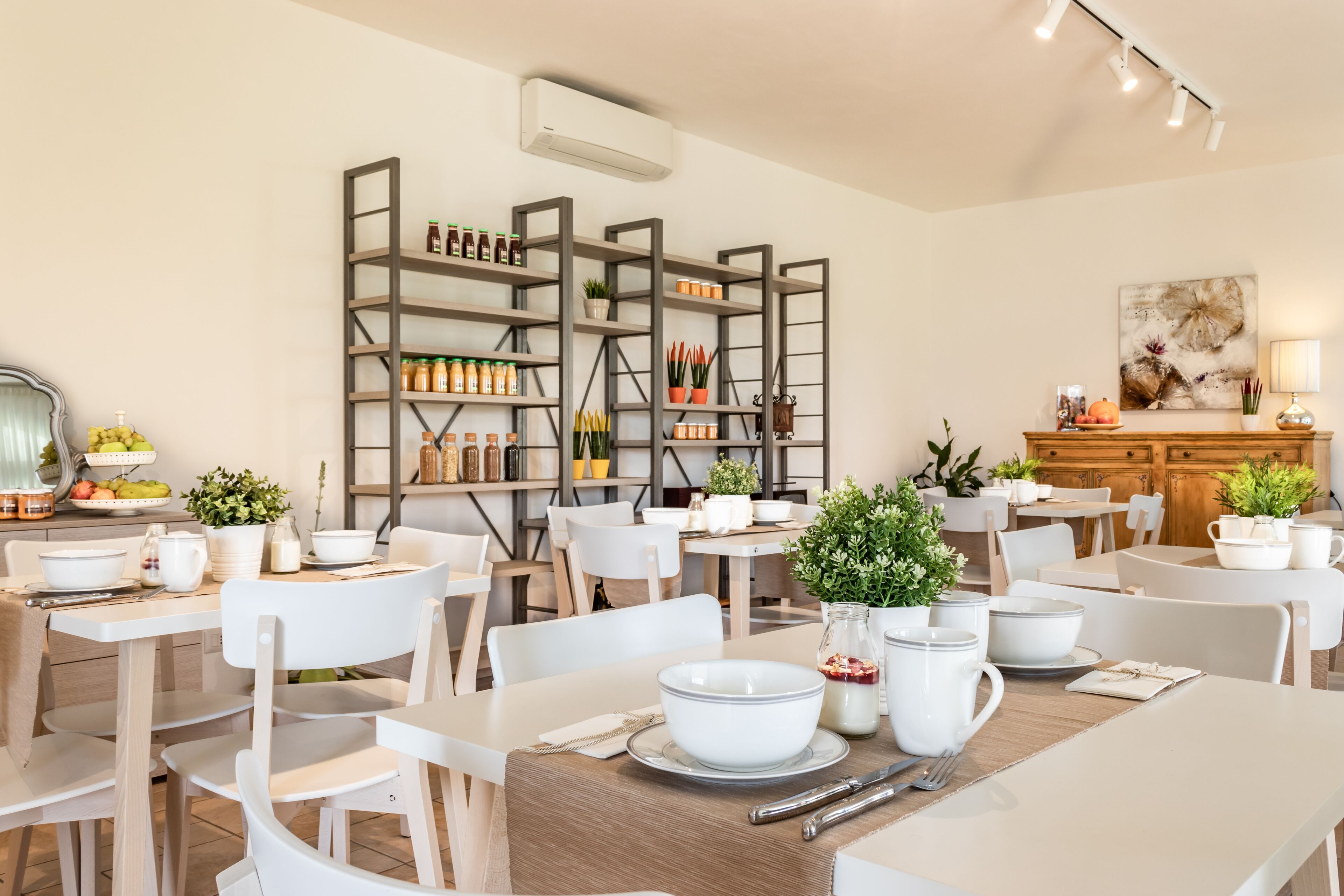 Ein Essbereich mit weißen Tischen und Stühlen, ein zentraler Holztisch mit einer weißen Schüssel und ein Regal mit Gläsern und Pflanzen im Hintergrund.