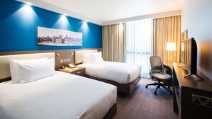 A hotel room with two beds, a desk, a chair, a TV, and a cityscape painting on the wall. - Hampton by Hilton Hamilton Park (Hamilton)