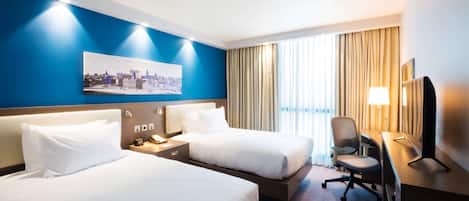 A hotel room with two beds, a desk, a chair, a TV, and a cityscape painting on the wall.