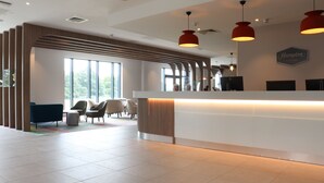 A modern hotel reception area with a white reception desk, red pendant lighting, and a large window offering a view of the outdoors. - Hampton by Hilton Hamilton Park (Hamilton)