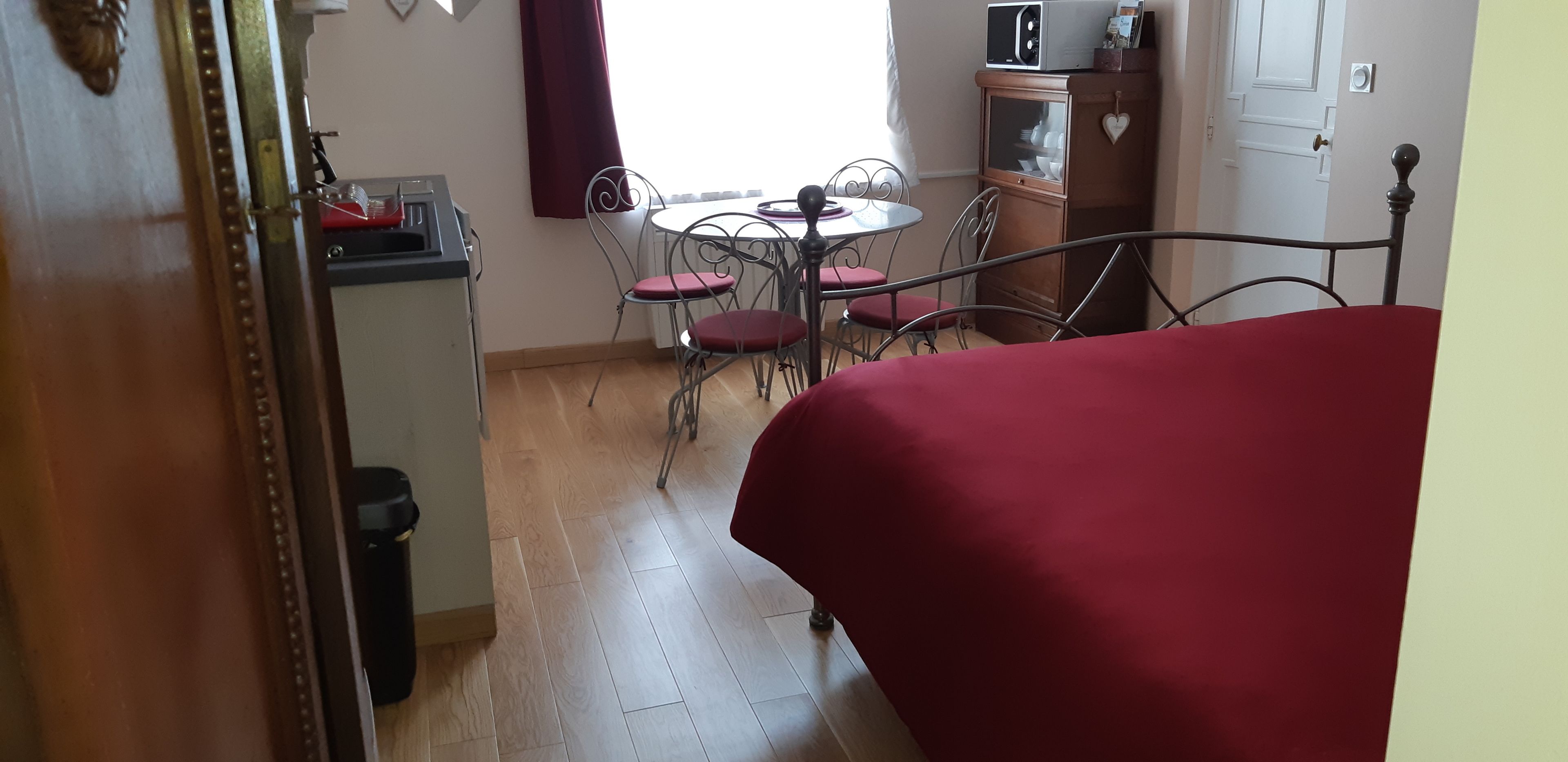 Un dormitorio con cama, una mesita, y una silla. Hay una ventana con cortinas, una puerta, y un armario con microondas.