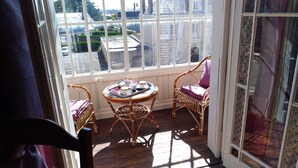 A sunlit room with a wooden floor, two wicker chairs, a small round table with a tray of items, and a view of buildings through large windows. - Chambres d'Hôtes Les Clefs du Bonheur (Chateau-Gontier)