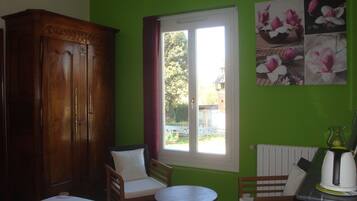 A room with a green wall, a wooden wardrobe, two chairs, a small round table, and a window with a view of the outside.