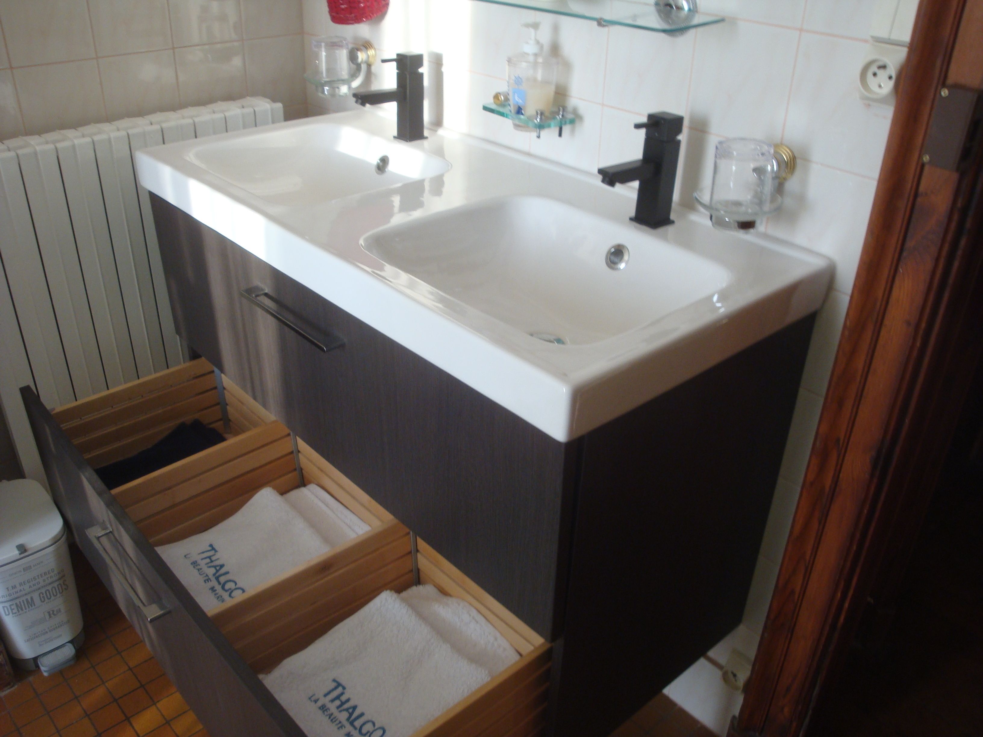 A bathroom with two sinks, a wooden drawer unit, and a glass shelf above the sinks.