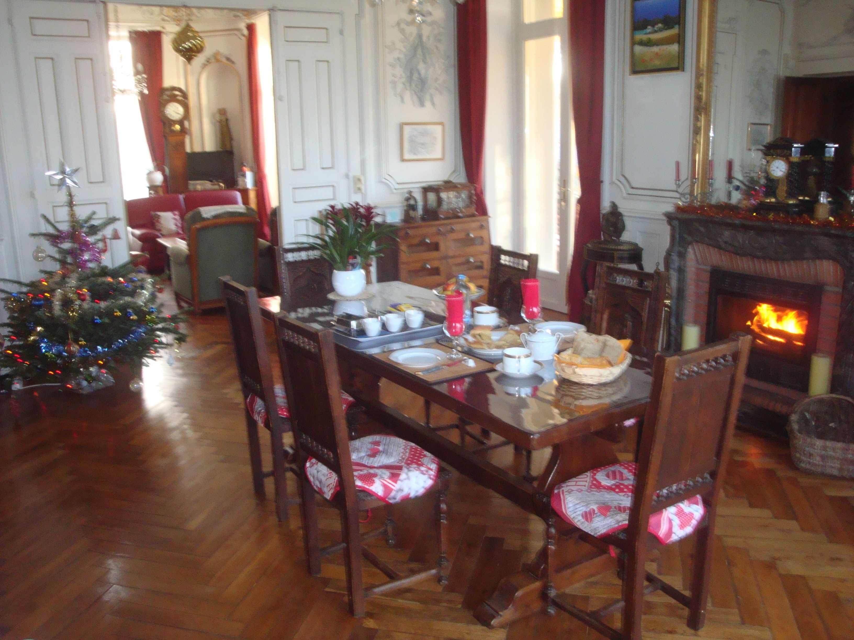 A dining area with a wooden table set for a meal, surrounded by chairs with patterned cushions. A Christmas tree with lights is visible in the foreground.