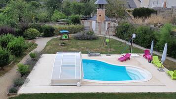Ein Schwimmbecken mit integriertem Whirlpool, umgeben von einer Terrasse mit Liegestühlen und einer kleinen Spielanlage.