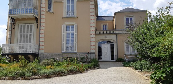 A two-story building with a balcony, a door with a decorative arch, and a gravel pathway leading to the entrance.