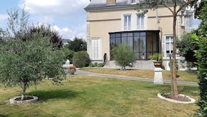 A well-maintained garden with a variety of trees and shrubs, a stone pathway, and a large conservatory attached to a building with a classic architectural style. - Chambres d'Hôtes Les Clefs du Bonheur (Chateau-Gontier)
