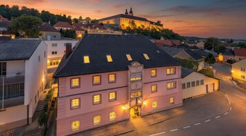 A pink building with multiple windows and a central entrance, surrounded by a cobblestone street and other residential buildings.