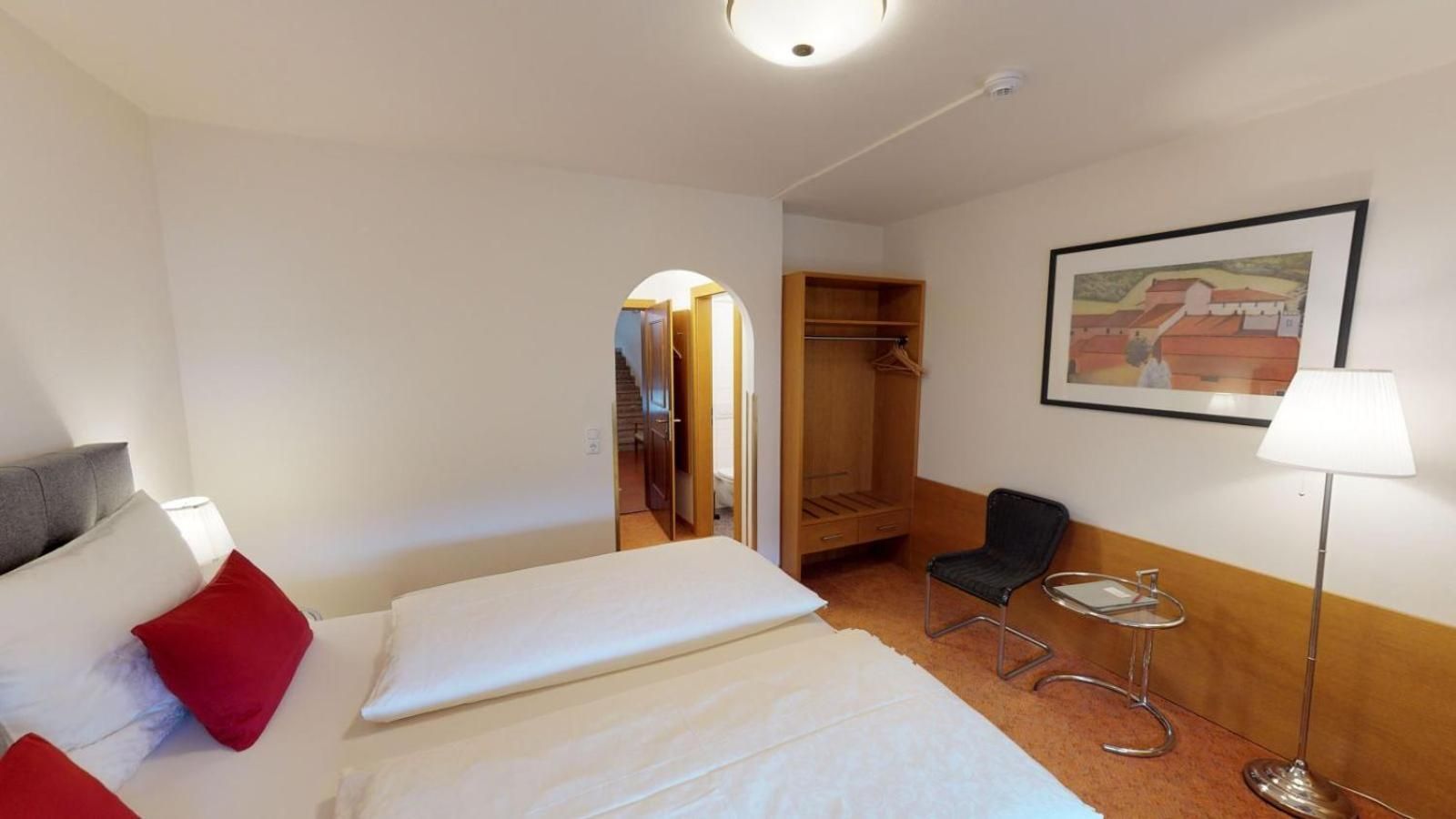 Ein Hotelzimmer mit Bett, Stuhl, Schreibtisch, Lampe, Kleiderschrank und einem Gemälde an der Wand.