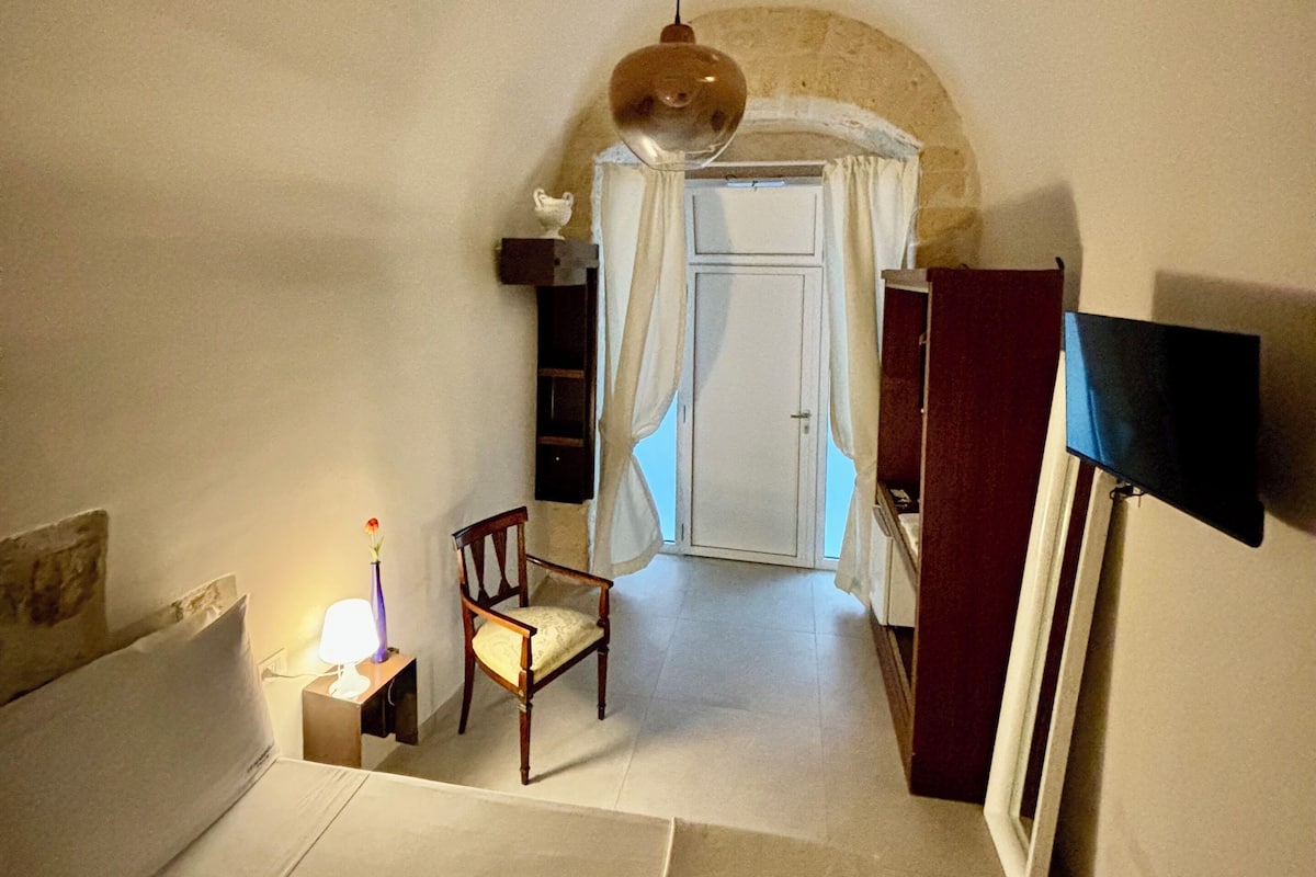 A narrow room with a bed, a chair, a small table with a lamp, a hanging light fixture, and a door leading to another room.