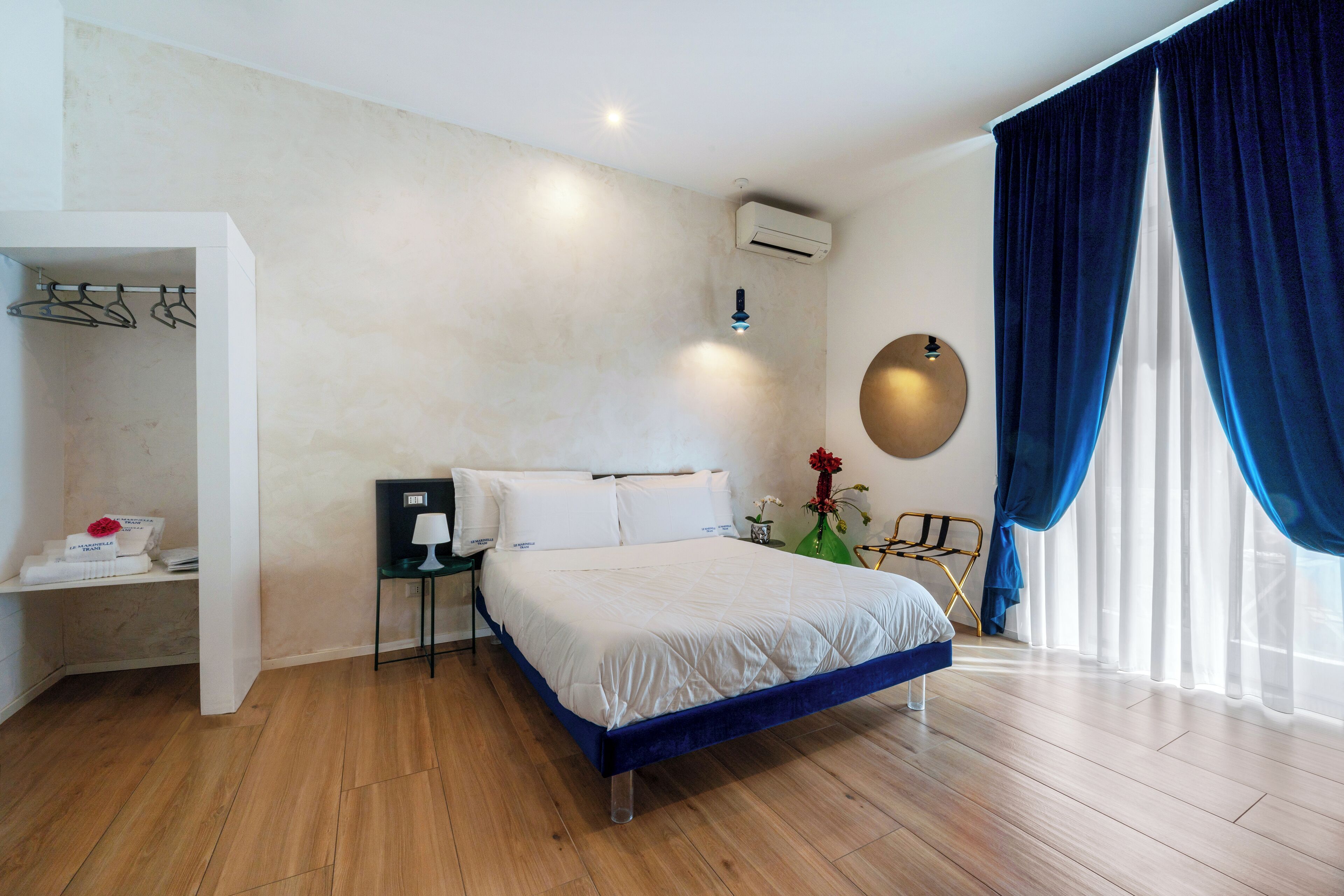 Una habitación de hotel moderna con piso de madera, una cama con cabecera azul, una mesita de noche con lámpara, aire acondicionado de pared y un amplio ventanal con cortinas azules.