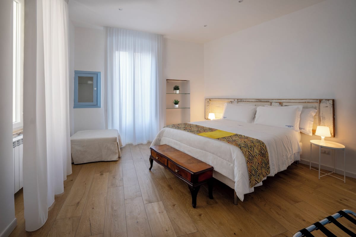 A bedroom with a wooden floor, a bed with white bedding, a bedside table with a lamp, a bench, and a window with curtains.