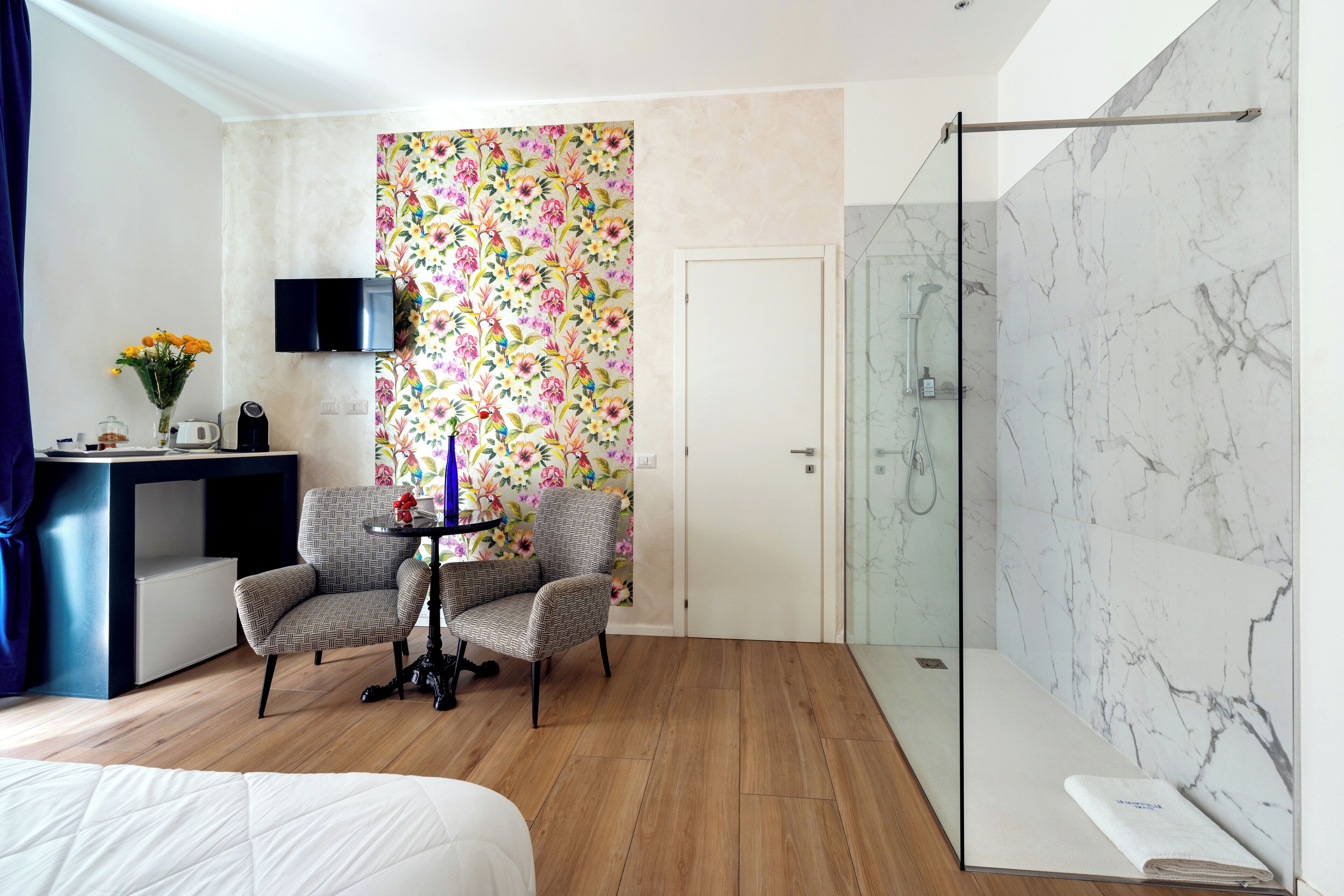 A hotel room with a bed, two armchairs, a small table with a vase of flowers, a TV, a shower with marble walls, and a door with a floral wallpaper.