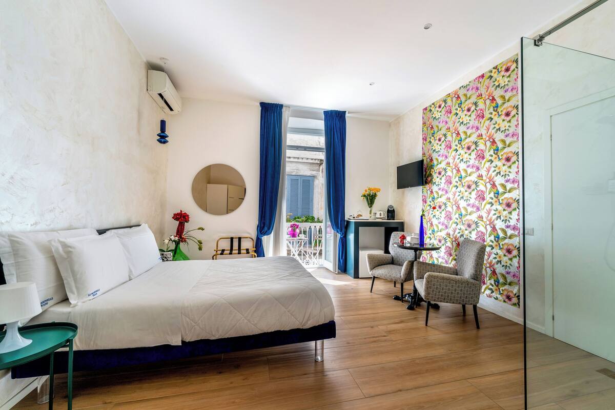 A hotel room with a bed, a desk, a chair, a TV, and a balcony with blue curtains.