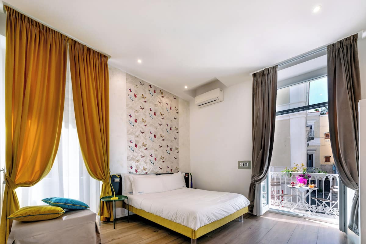 A bedroom with a bed, a chair, a small table with a plant, and a balcony with a railing.