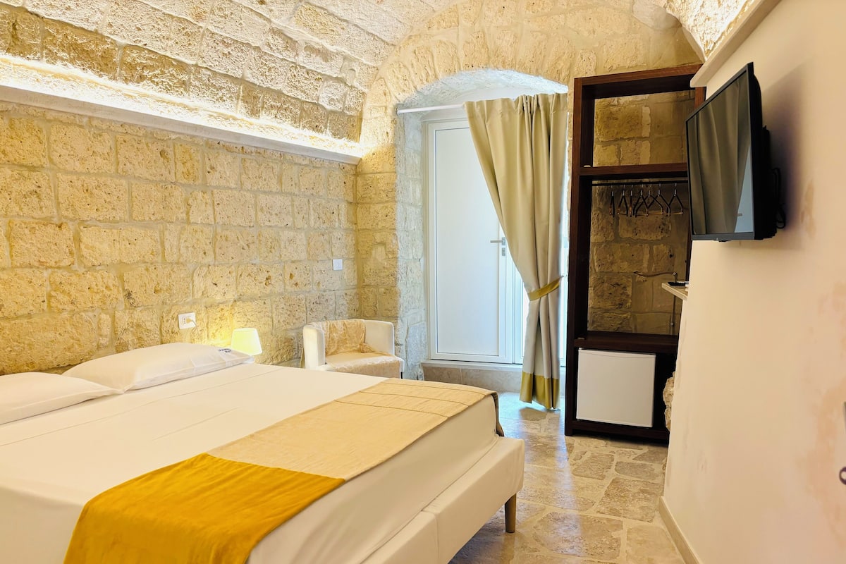 A bedroom with a large bed, a chair, a TV mounted on the wall, and a stone wall.