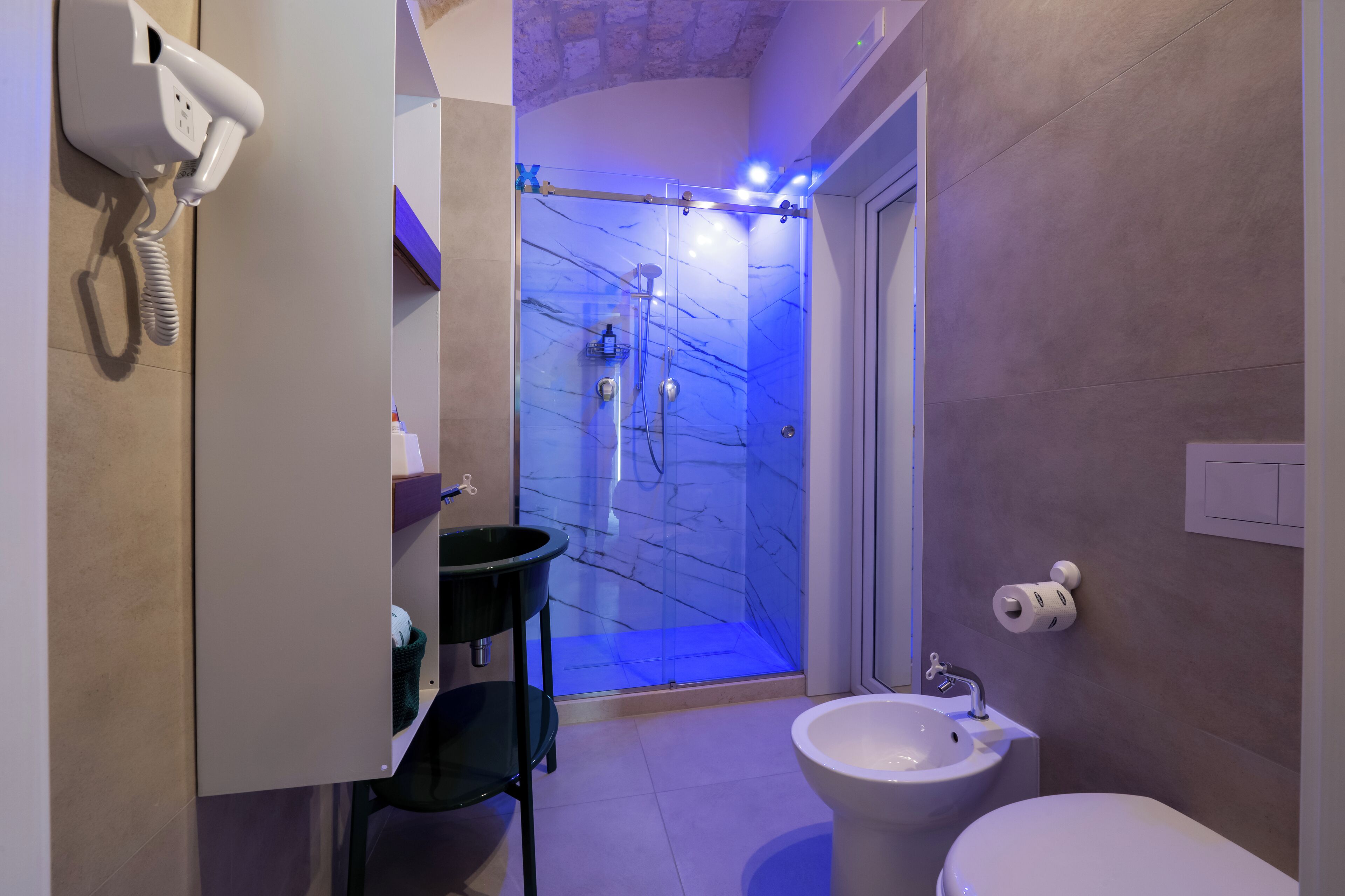 Un baño con ducha de mármol, lavamanos blanco, inodoro y secador de pelo montado en la pared.