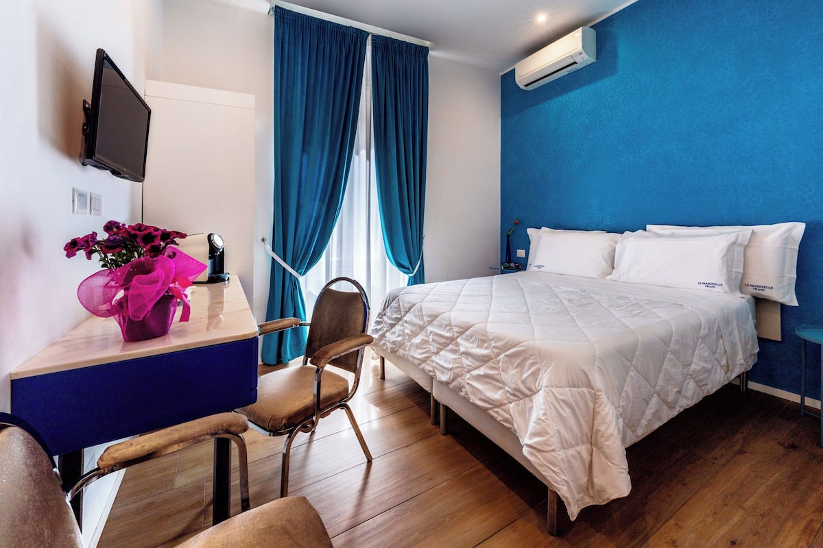 A hotel room with a bed, a desk with a chair, a TV mounted on the wall, and a window with blue curtains.
