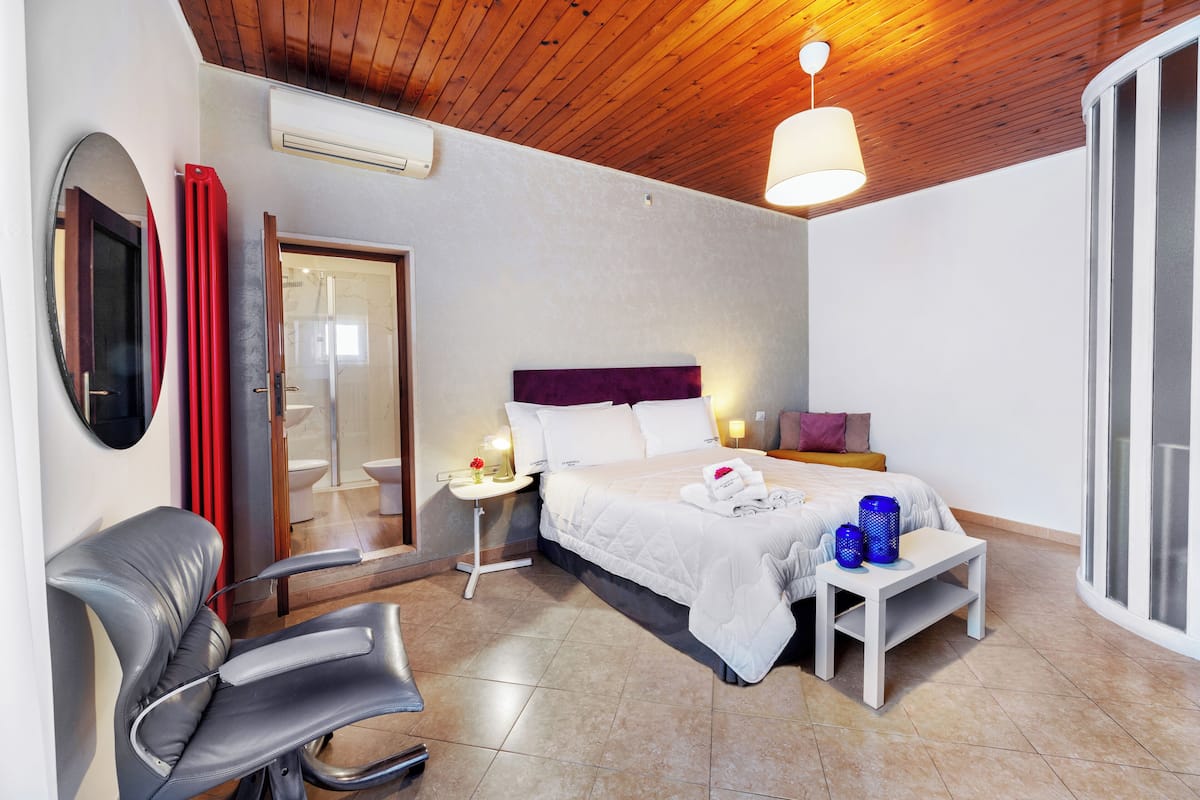 A hotel room with a bed, a chair, a bedside table, and a window.